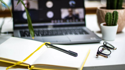 An open notebook with a pen laying on top. A blurred laptop screen in the background shows an ongoing online meeting.