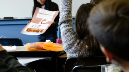 A school child holds their hand up as another child stands in the front of the classroom whilst presenting