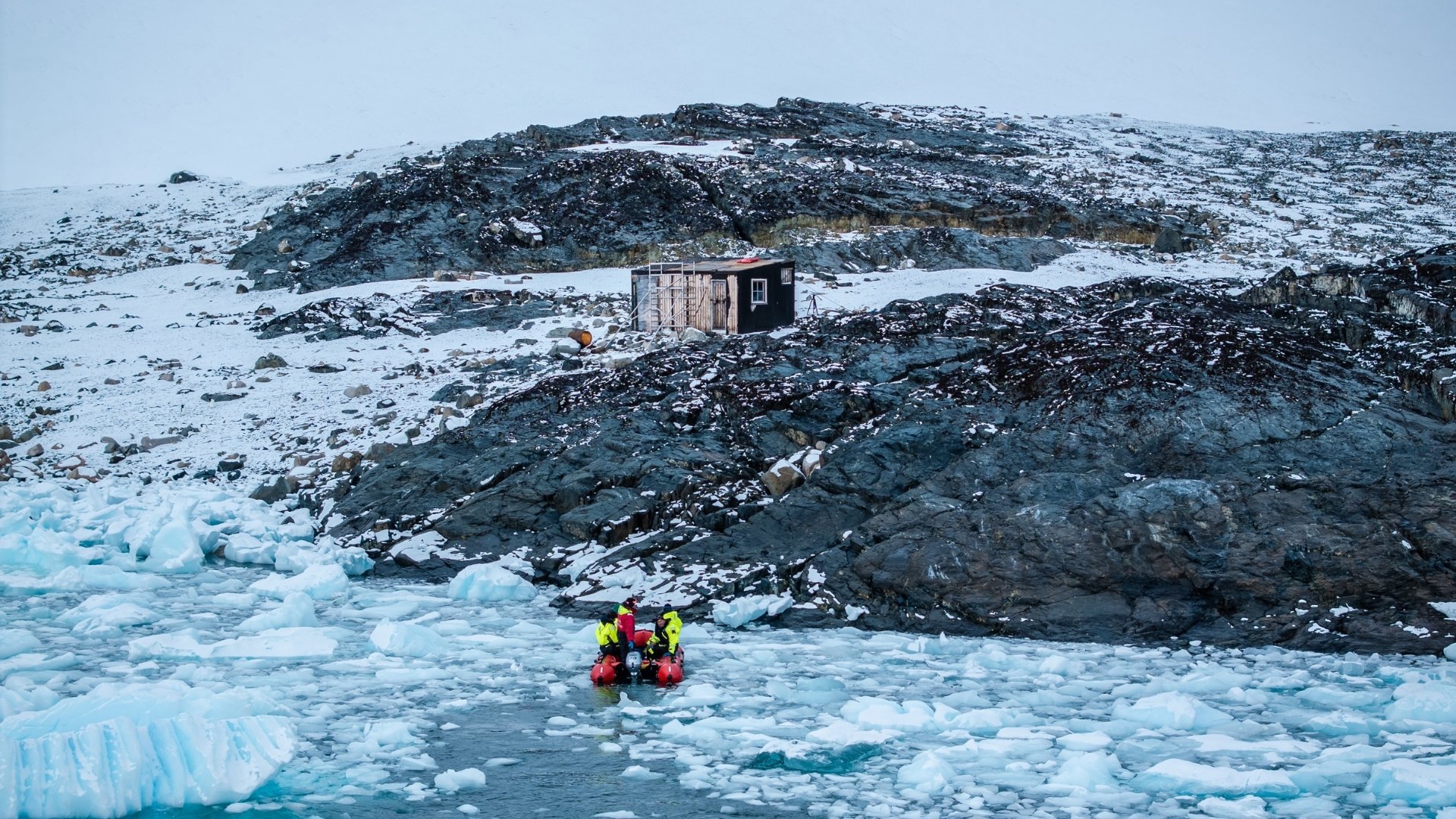 Two people on a small boat aproaching a hut on a snow covered island.