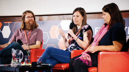 Three people sititng in comfortable chairs on a stage, with the person in the middle holding a microphone and speaking.
