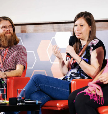 Three people sititng in comfortable chairs on a stage, with the person in the middle holding a microphone and speaking.