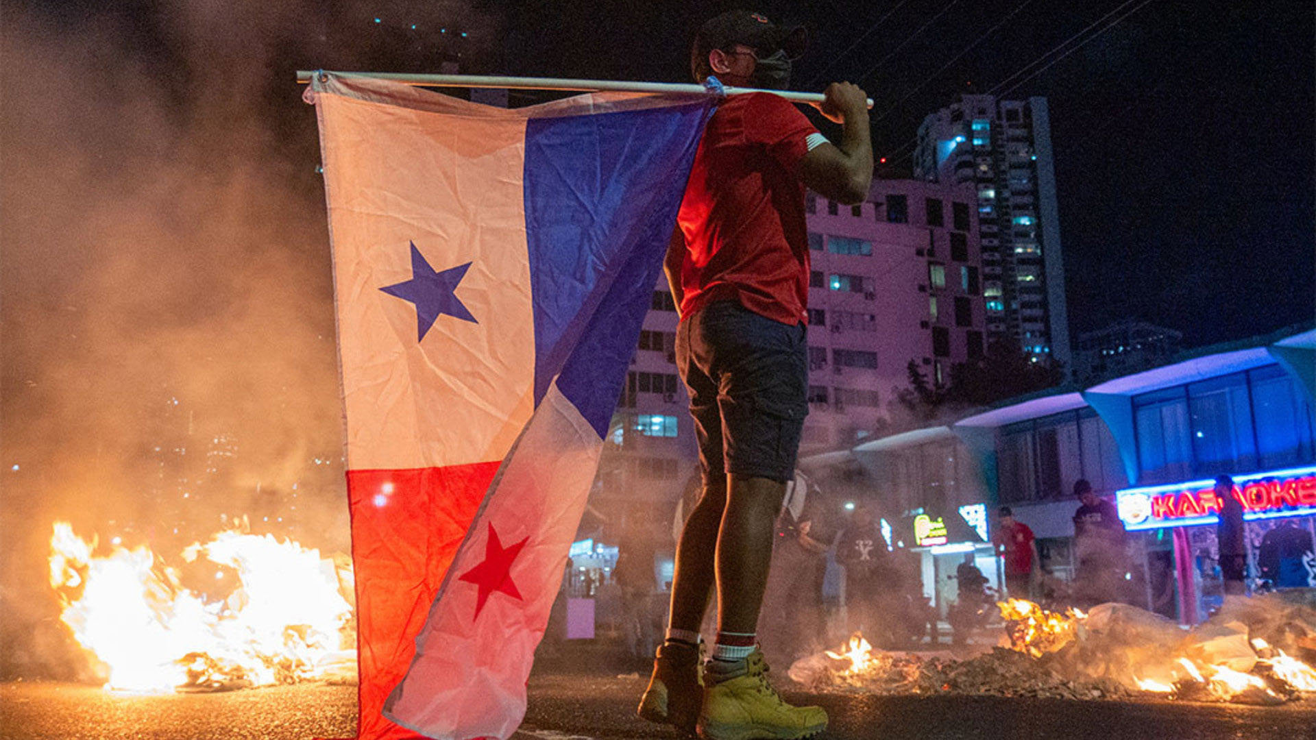Person holding a Panama flag near burning debris during a night-time protest in a city street.