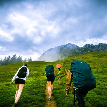 Four people with large backpacks are walking up a green hill towards a rocky outcrop. The clouds are low and are forming a mist at the top of the hill towards where they are heading