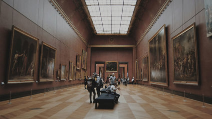 People looking at paintings hung on walls in a gallery hall of Musée du Louvre, Paris, France.