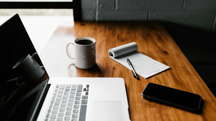 An open laptop, coffee mug containing coffee, notebook, pen and mobile phone on a desk.