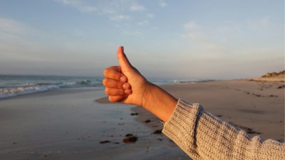 Hand with thumb up against beach background.
