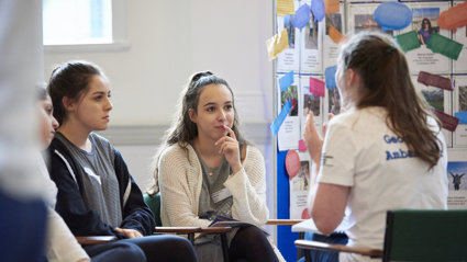 Two students listening attentively to a third person who's wearing a T-shirt with the text Geography Ambassador.
