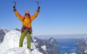 Expeditioner in winter gear holding up two pickaxes while standing on the summit of a snow covered mountain.