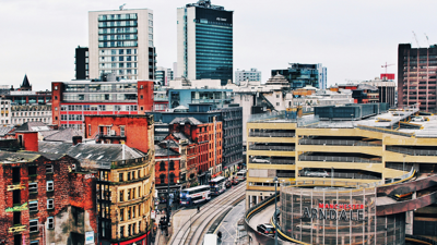 A rooftop view of Manchester city centre. Tall red brick buildings stand in the shadows of glass skyscrapers. train tracts cut through the urban centre.