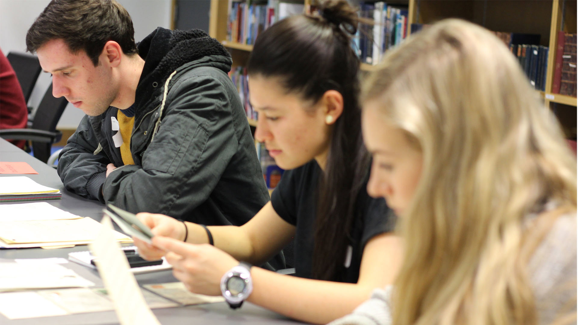 Three people sat at a table in a library, looking at paperwork.