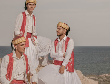 Three men in traditional clothing sitting on a cliff next to the sea.