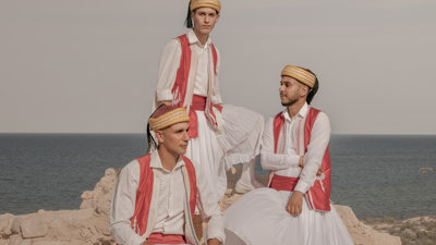 Three men in traditional clothing sitting on a cliff next to the sea.
