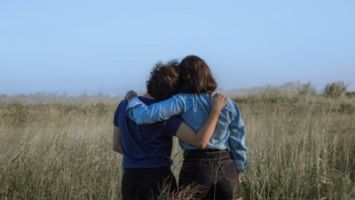 Two people as seen from behind standing in a spacious meadow with an arm around each other's shoulder.