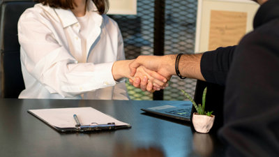 Two people sit across from each other at a table whilst shaking hands. There is a clip board and pen on the table to their left and laptop on the table to their right.