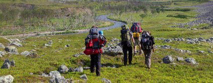 Four people with large rucksacks and camping equipment walking beside a meandering river. There are trees on the bank of the river and piles of rocks across the grass where they are walking.