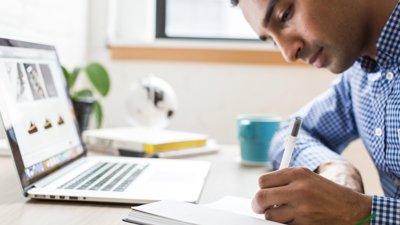 Person writing notes looking at laptop