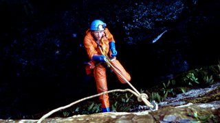 Man caving in his full gear, holding a rope while hanging above a cave.