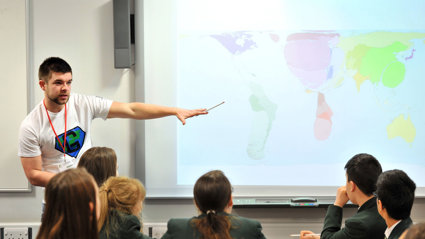 Group of students watching a teacher, who's pointing at a world map on a screen. 