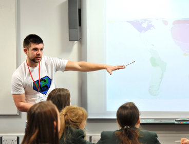 Group of students watching a teacher, who's pointing at a world map on a screen. 