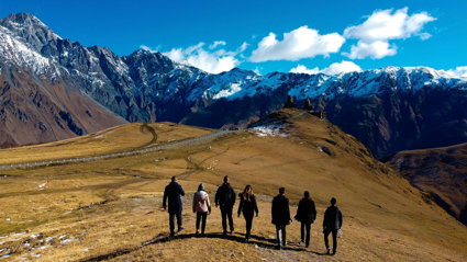 A group of seven people on a brown field facing snow-covered mountains on a sunny day.