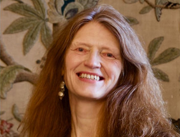 Person with long brown hair wearing a black blazer against a floral wallpaper background.