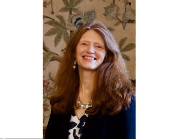 Person with long brown hair wearing a black blazer against a floral wallpaper background.