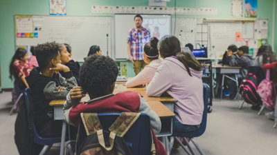 A teacher standing at the front of a classroom with students sat at the desks.