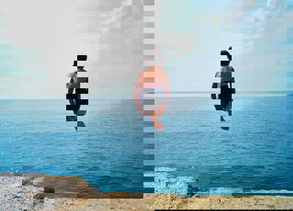 Person curled up, diving off a cliff into an blue ocean