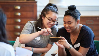 Two students sitting on chairs and smiling while looking at a phone together.