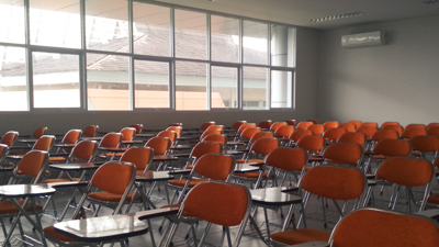 Orange chairs neatly fill an empty class room. large square panel windows on the left of the room let in beams of light.