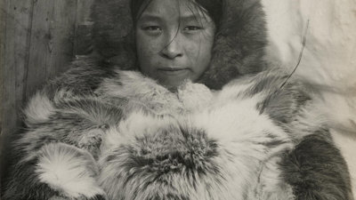Black and white photo of an Inuit woman called Aqattaq. Aqattaq stares forward with a nutural expression wearing a fur top and fur hood.