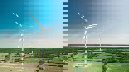 A view of two white wind turbines, located in flat countryside with fields and forest