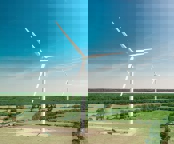 A view of two white wind turbines, located in flat countryside with fields and forest