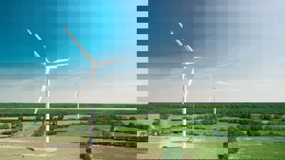 A view of two white wind turbines, located in flat countryside with fields and forest