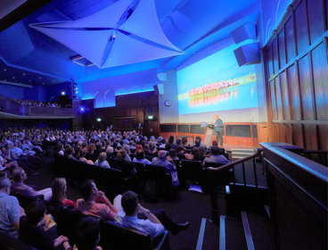 Audience members attending a lecture. One speaker is standing in front of a projection screen on a stage presenting.