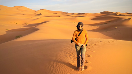 Person with camera wearing orange jacket in the middle of an sandy orange Morocco desert.