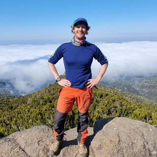 Conor Diffin standing on a mountain ledge overlooking a forested valley.