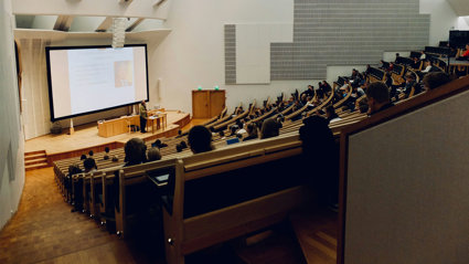 Lecture theatre with students sitting in staggered rows of seats. The rows are faced towards a stage with someone giving a presentation in front of a screen. 