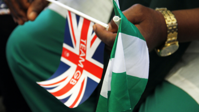 A person holds both the red, white and blue union jack alongside the white and grey Nigerian flag. Their shirt is white and toursers are green, matching the flag of Nigeria.