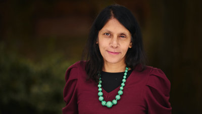 Portrait photo of Halima Begum smiling subtly, wearing a large, turquoise beaded necklace and maroon coloured top.