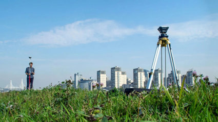 Surveying tool set up on a tripod in a field. There are skyscrapers in the background below a clear sky.