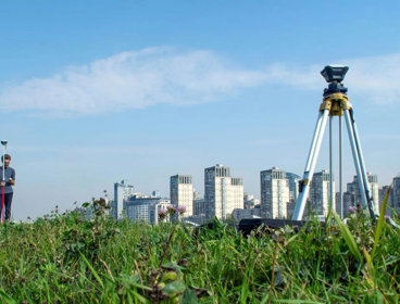 Surveying tool set up on a tripod in a field. There are skyscrapers in the background below a clear sky. 