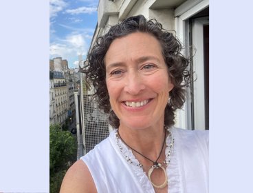 Person with curly hair wearing a white top standing on a balcony overlooking a cityscape with buildings and trees under a blue sky.