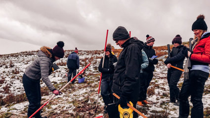 A group of seven secondary school pupils holding ranging poles standing on a snowy hillside.