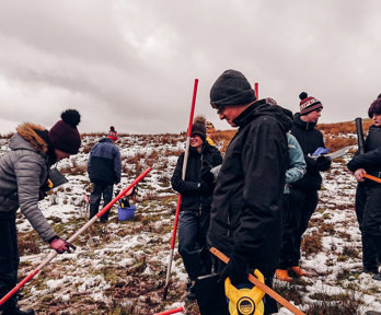 A group of seven secondary school pupils holding ranging poles standing on a snowy hillside.