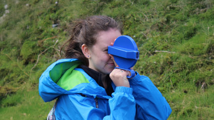 Child in blue jacket looking through a small handheld viewer outdoors on a grassy hillside.