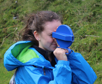 Child in blue jacket looking through a small handheld viewer outdoors on a grassy hillside.