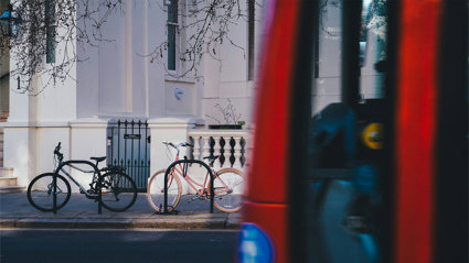 Two bicycles locked to racks outside a white townhouse, with a red double-decker bus passing in the foreground.