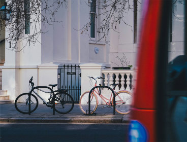 Two bicycles locked to racks outside a white townhouse, with a red double-decker bus passing in the foreground.