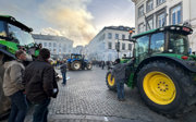 Farmers protest in Brussels with tractors blocking a cobbled street, smoke rising, and people holding banners in front of historic buildings.